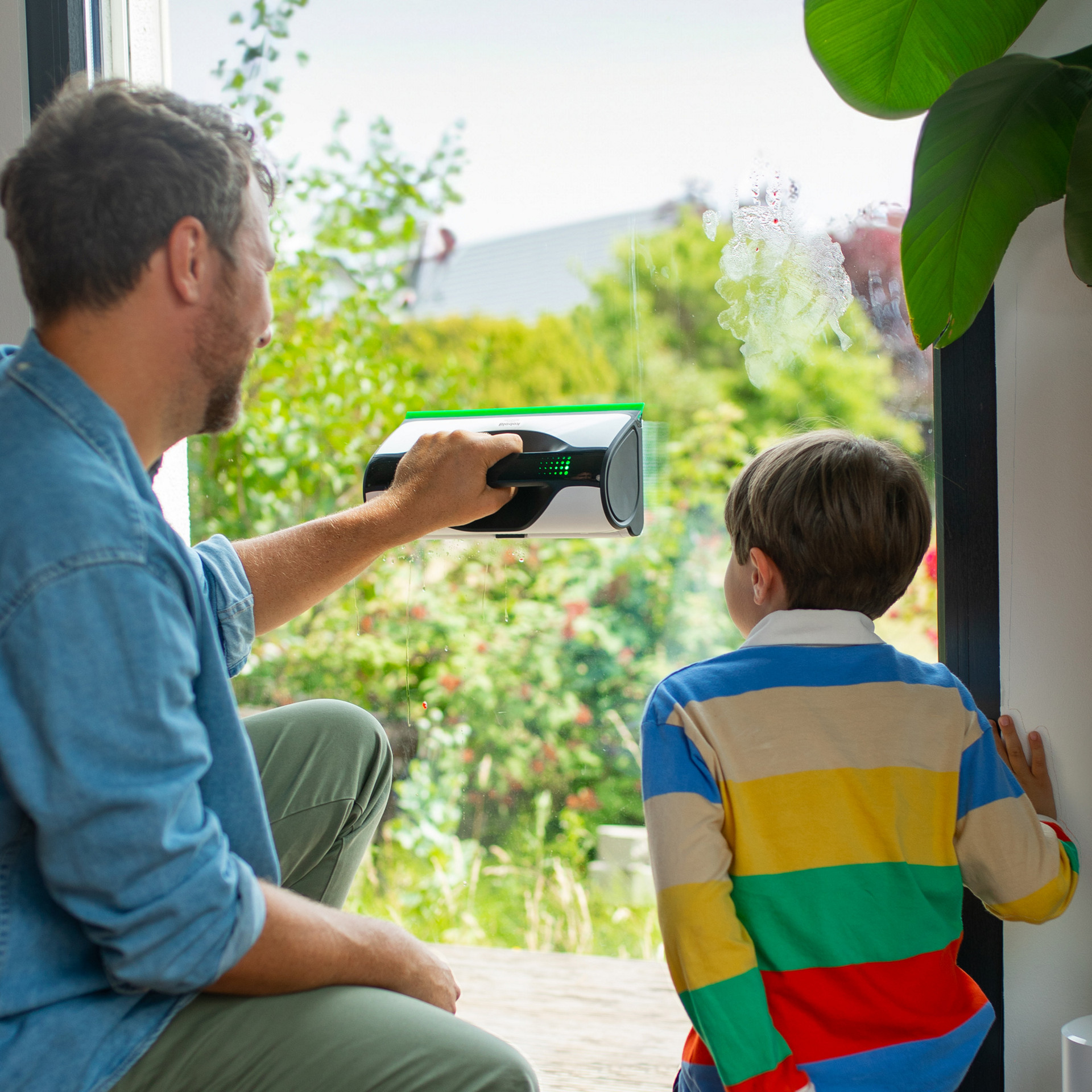 Erwachsener reinigt Fenster mit Kobold VG100+ Flächenreiniger, Kind schaut zu, nahe offener Tür und Pflanzen.