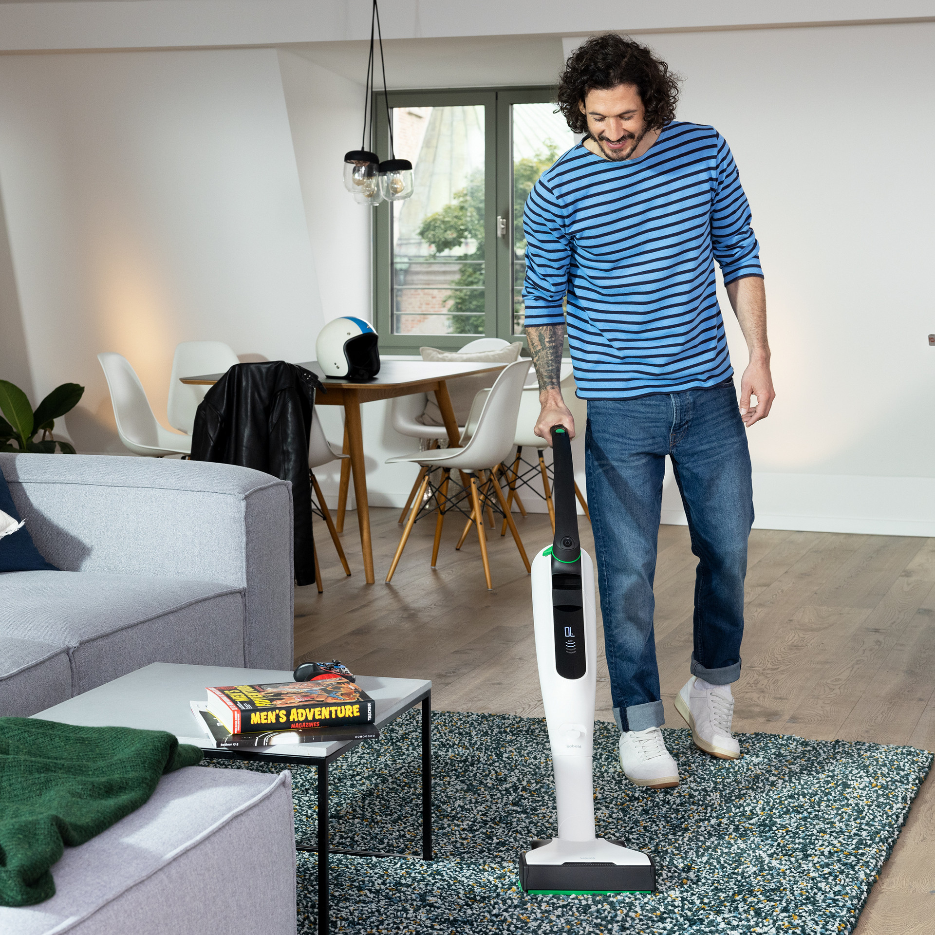 Osoba odkurza dywan w salonie za pomocą pionowego odkurzacza bezprzewodowego Kobold VK7 w nowoczesnym, jasnym wnętrzu.
