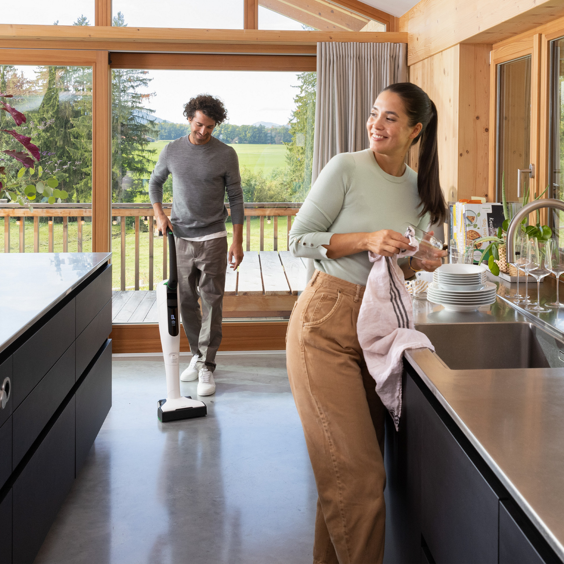 A person cleaning with a Kobold VK7 in a modern kitchen with wooden accents and large windows.