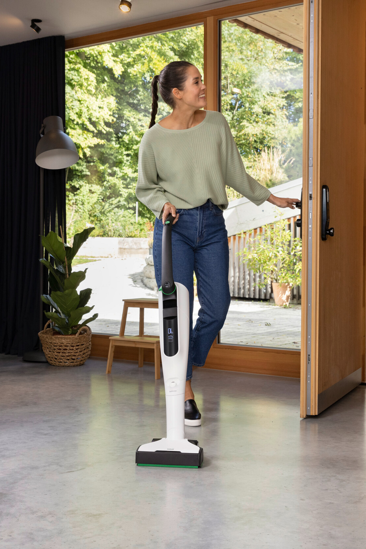 Frau nutzt einen Kobold VK7 in einem modernen Zuhause, während ein Mann mit einem Fahrrad hereinkommt.