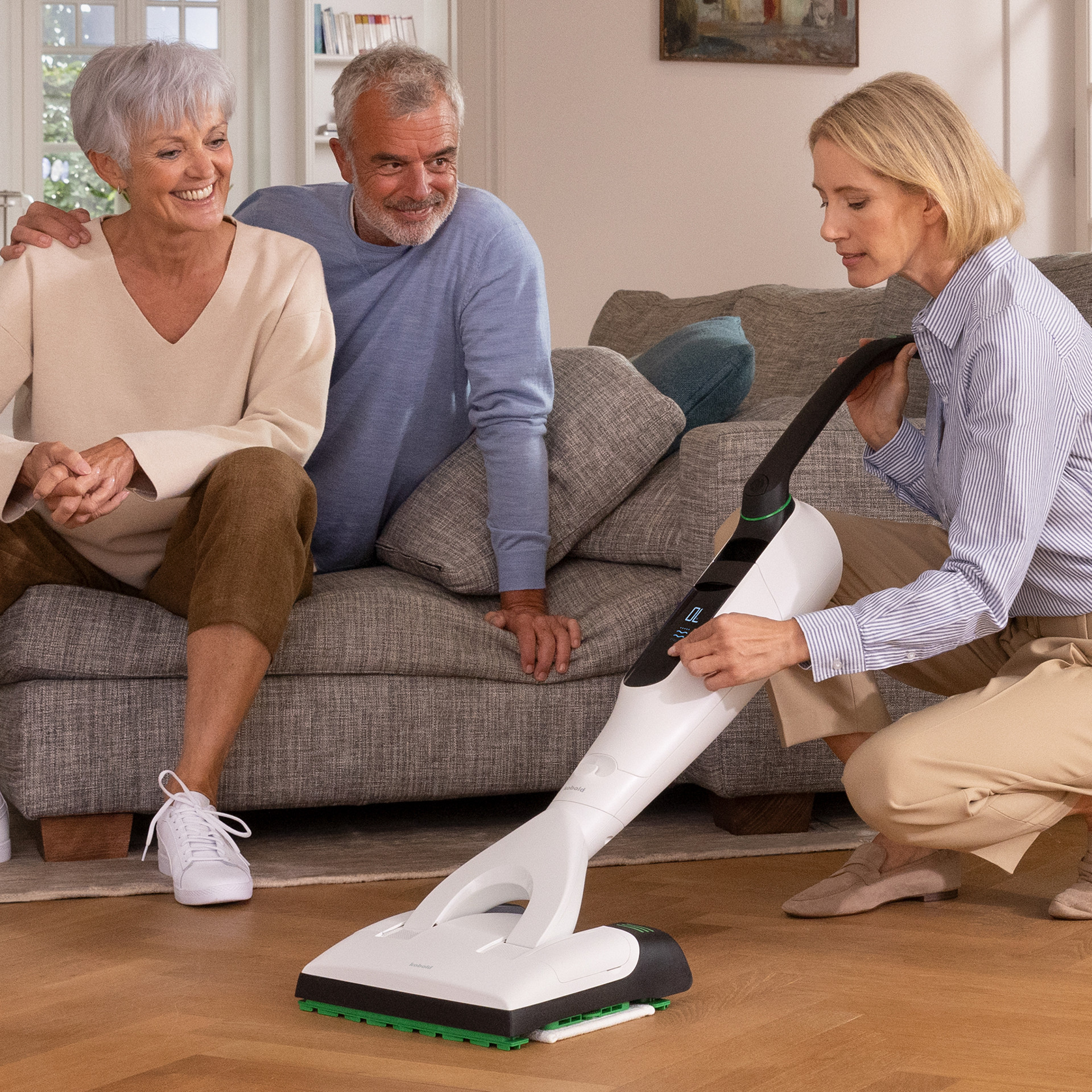 Frau demonstriert den VK7 Akku-Staubsauger SP7 Saugwischer auf Parkettboden im Wohnzimmer, zwei Personen sitzen auf dem Sofa.