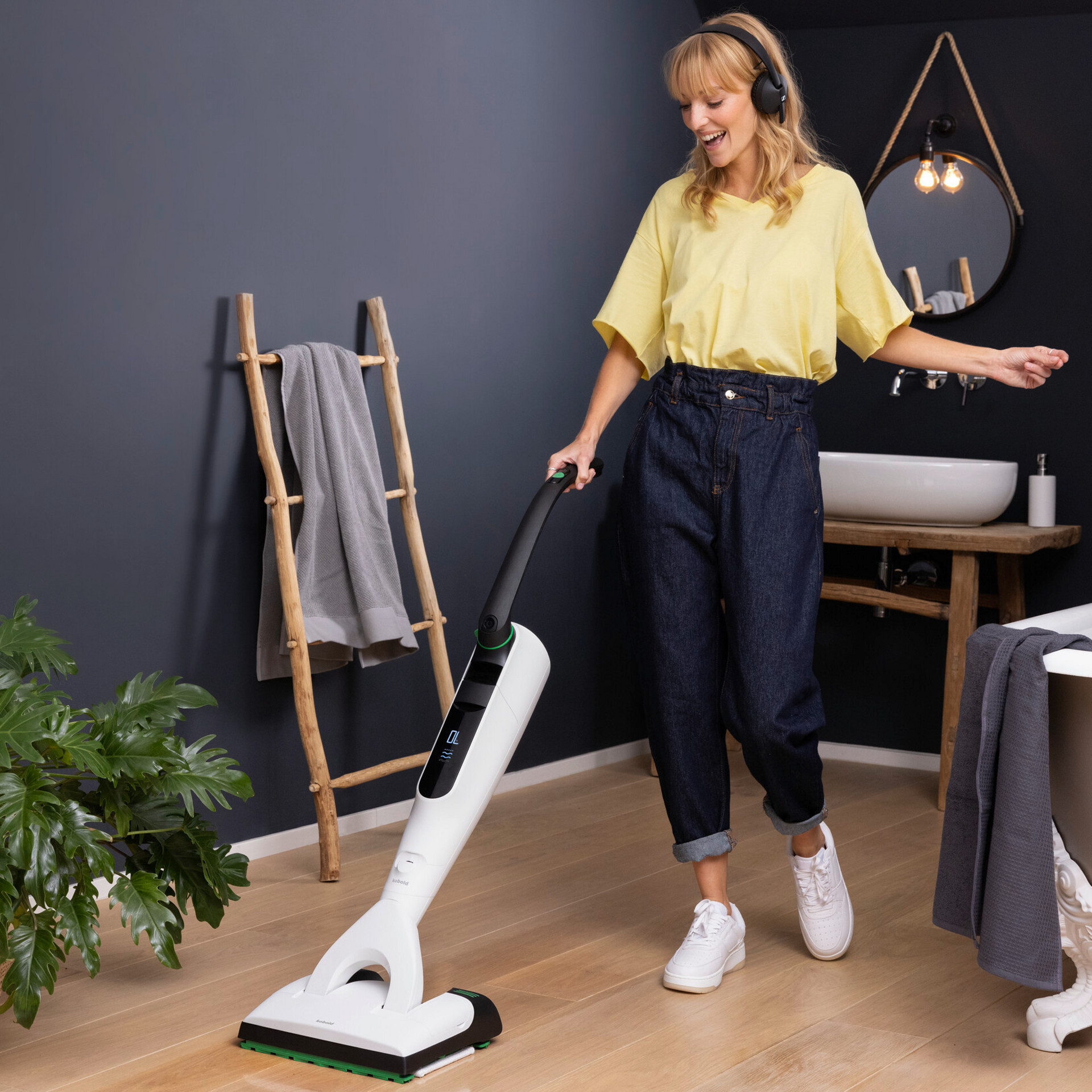 Eine Person reinigt den Boden eines modernen Badezimmers mit dem Kobold VK7, Holzdetails und grünen Pflanzen.