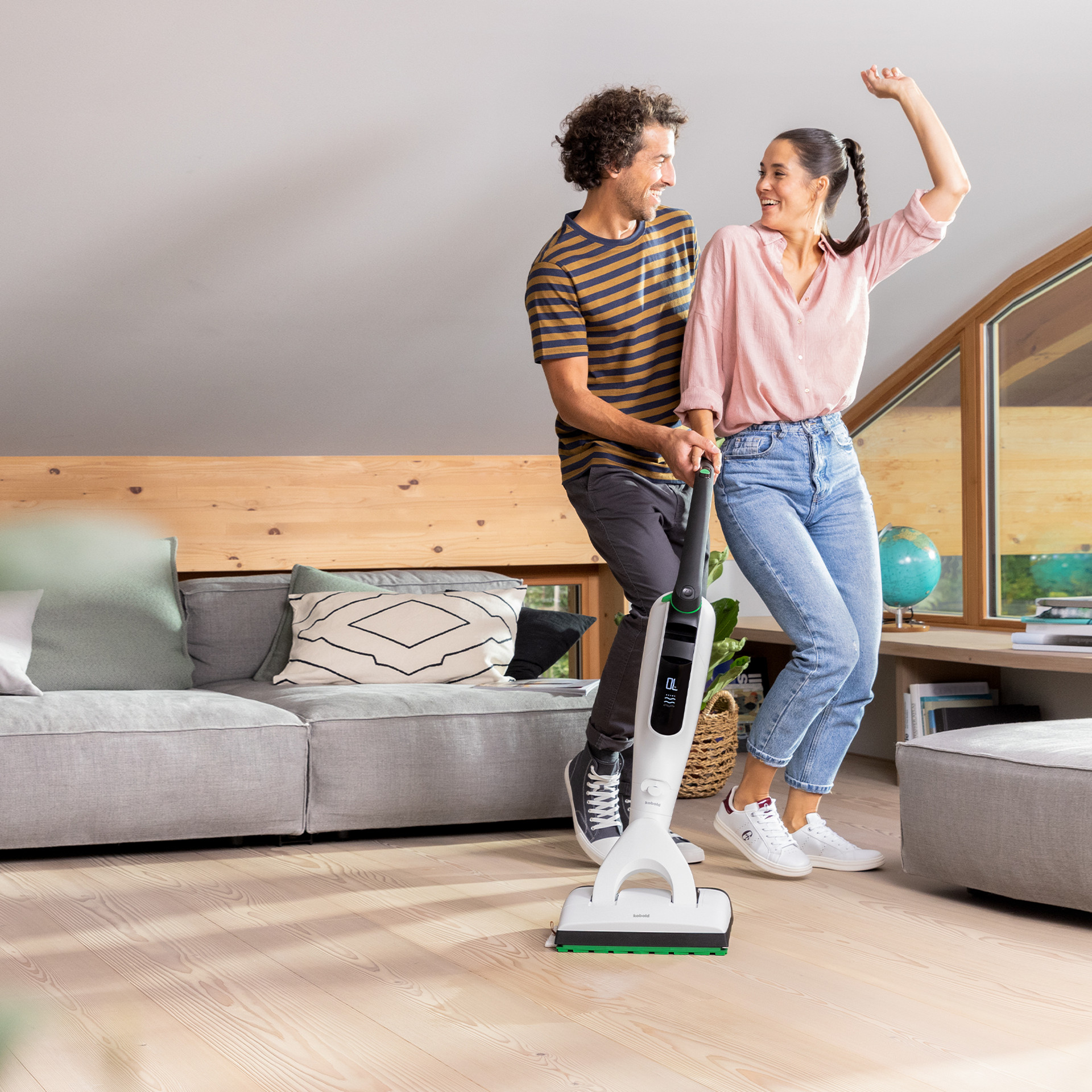 Zwei Personen reinigen ein Wohnzimmer mit einem Kobold VK7 vor einem modernen Sofa und großen Fenstern.