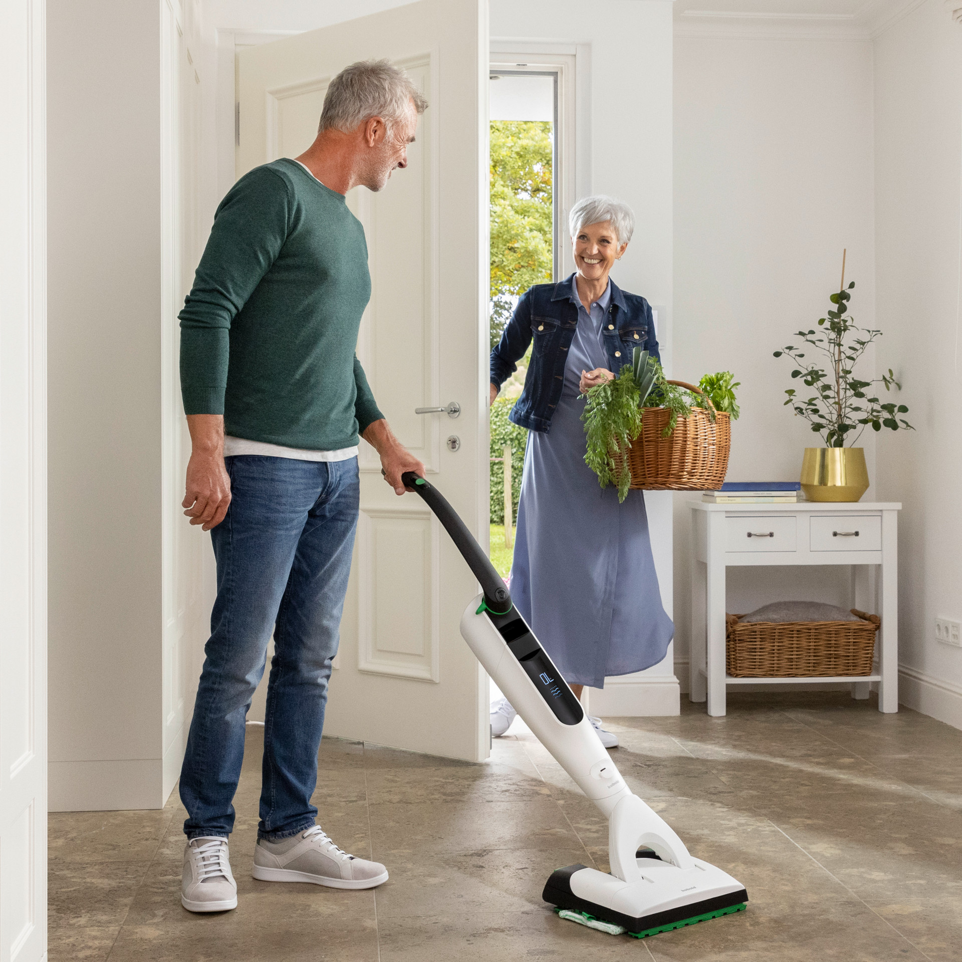 Man cleaning the floor of a bright entrance with a Kobold VK7 in front of a woman carrying a basket of fresh greens.
