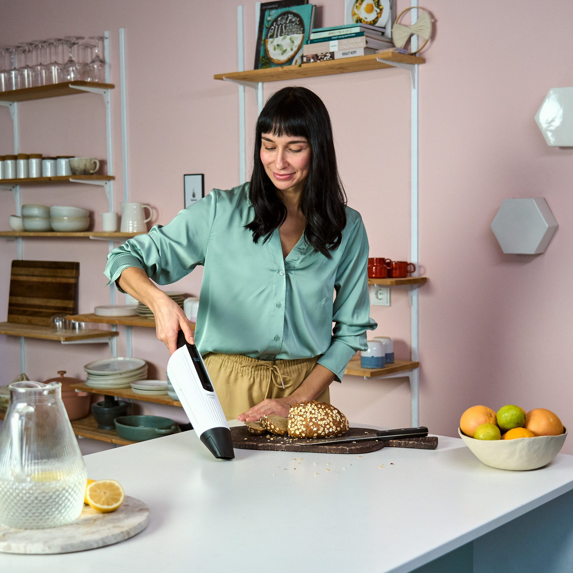 Person nutzt den Kobold VC100 Akkusauger, um Krümel von einer Küchenarbeitsplatte neben einem Brot zu entfernen.