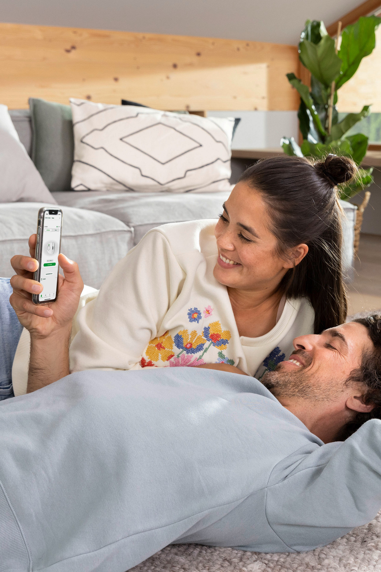 Couple relaxing on carpet while controlling the Kobold VR7 robot vacuum cleaner with a smartphone in a bright living room.