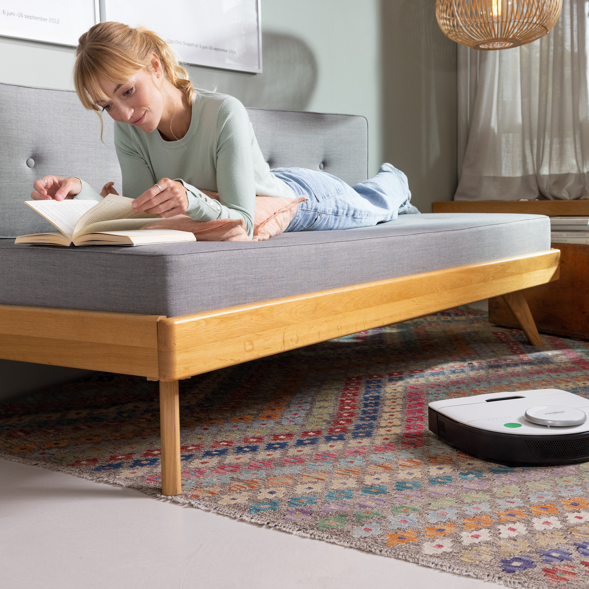 Kobold VR300 Saugroboter reinigt einen gemusterten Teppich in einem hellen Wohnzimmer, während eine Person auf dem Sofa liest.