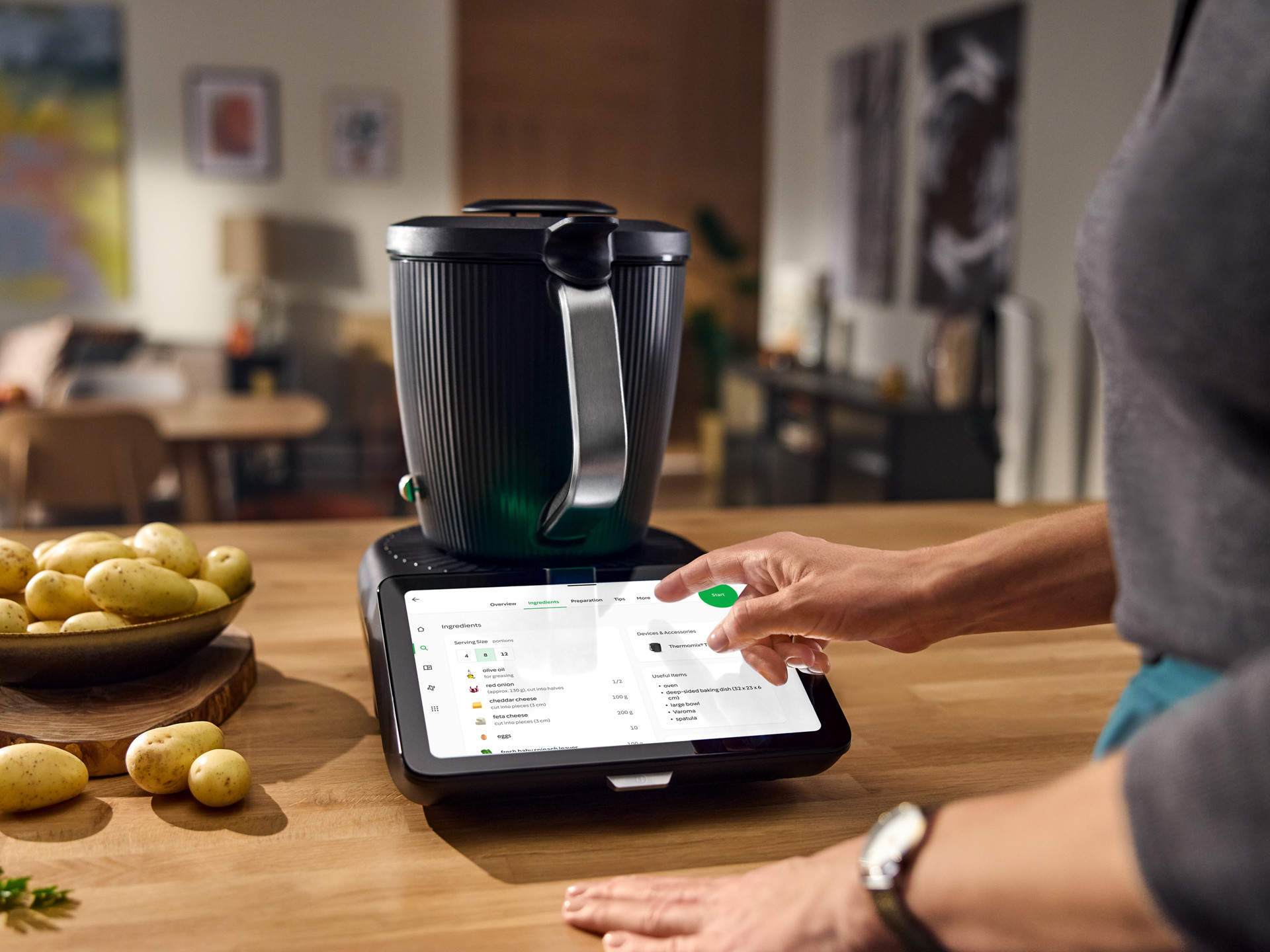 Persona seleziona una ricetta su Bimby TM7 touchscreen in cucina moderna con una ciotola di patate vicina.