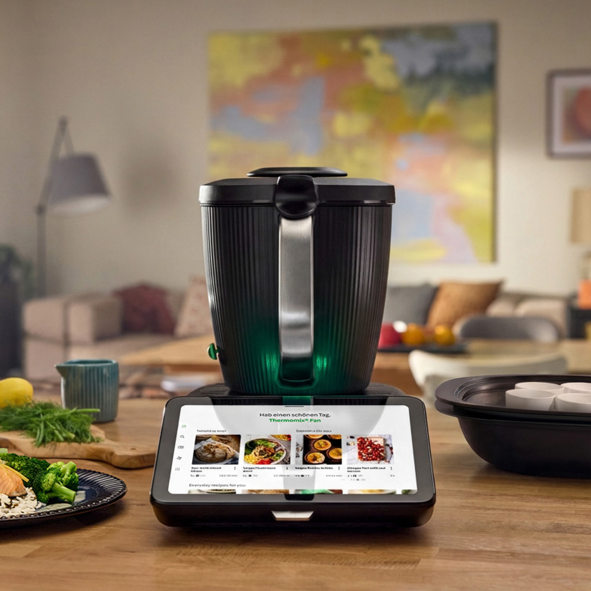 Thermomix® TM7 mit eingeschaltetem Display auf Küchenplatte, umgeben von Speisen und Varoma® mit Förmchen, hinten Wohnzimmer