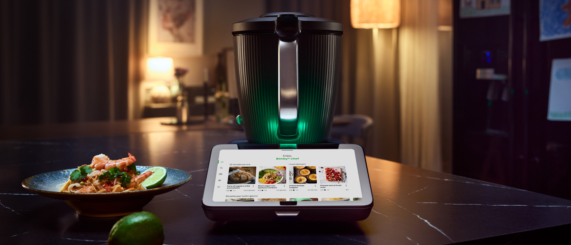 Bimby TM7 su piano cucina con display touch, piatto pronto, lime e peperoncini in primo piano.