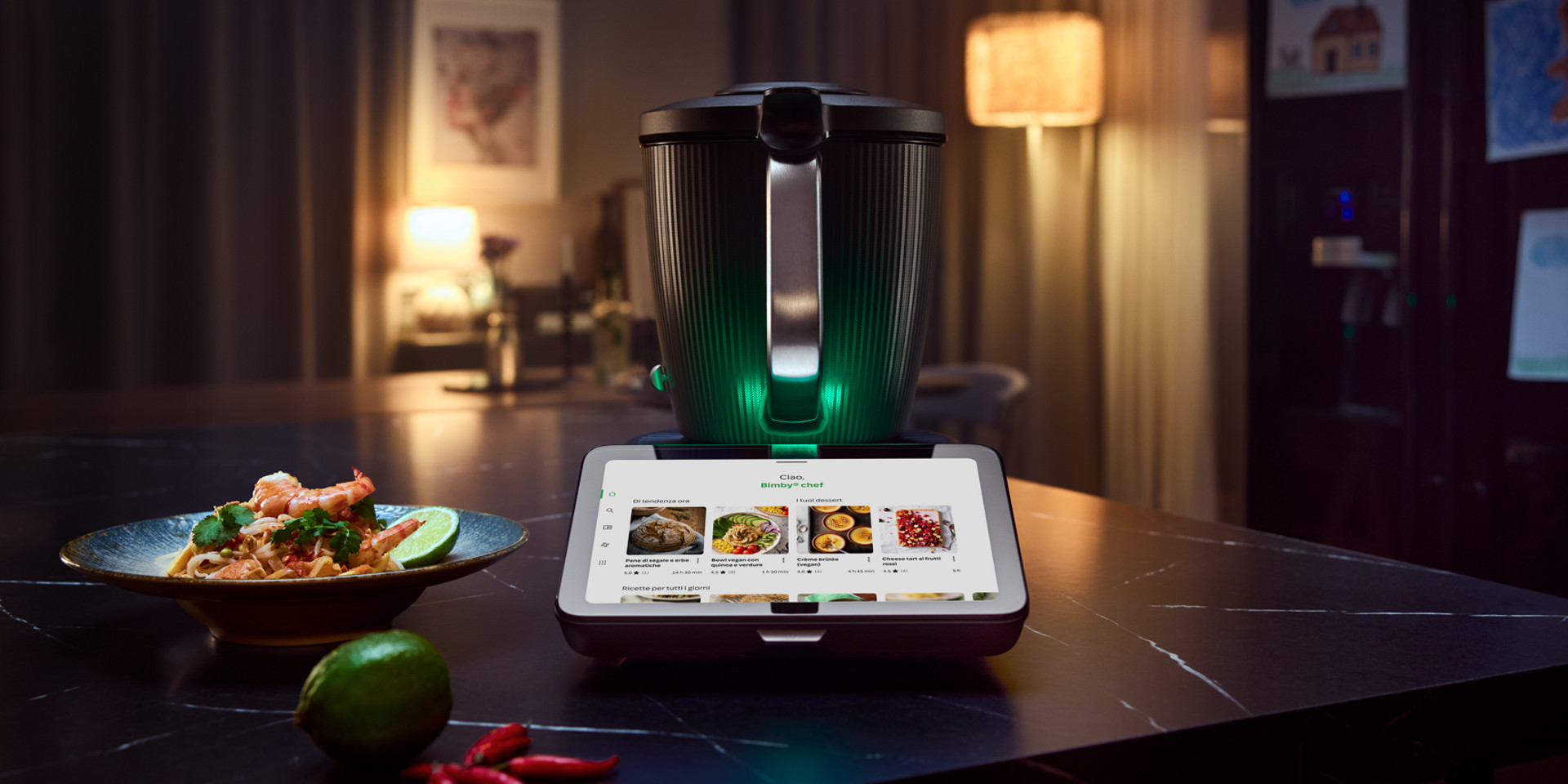 Bimby TM7 su piano cucina con display touch, piatto pronto, lime e peperoncini in primo piano.