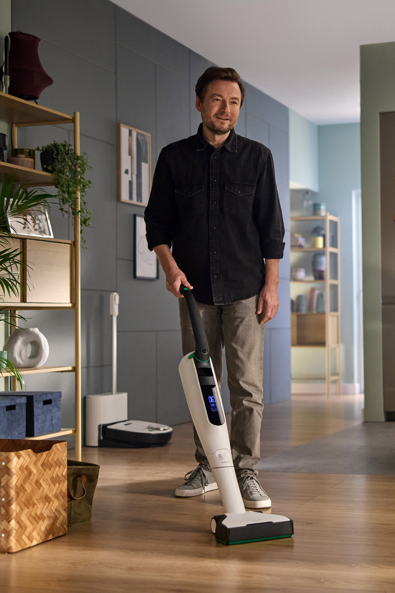 A man uses a Kobold VK7 in a living room while a Kobold VR7 and Thermomix® TM7 are visible in the background.