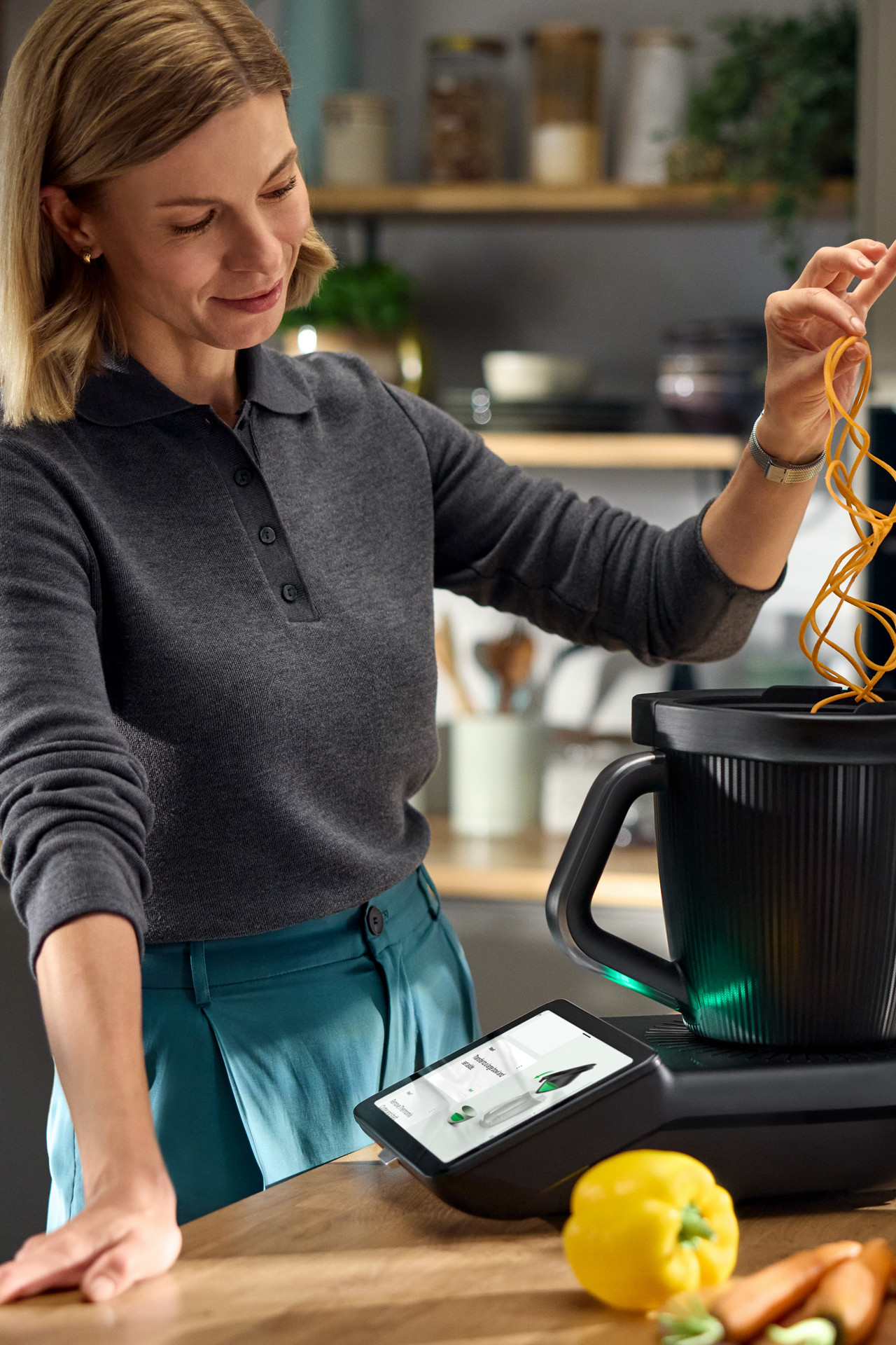 Frau benutzt Thermomix® TM7 mit spiralförmigem Gemüse in einer modernen Küche, umgeben von frischem Gemüse.