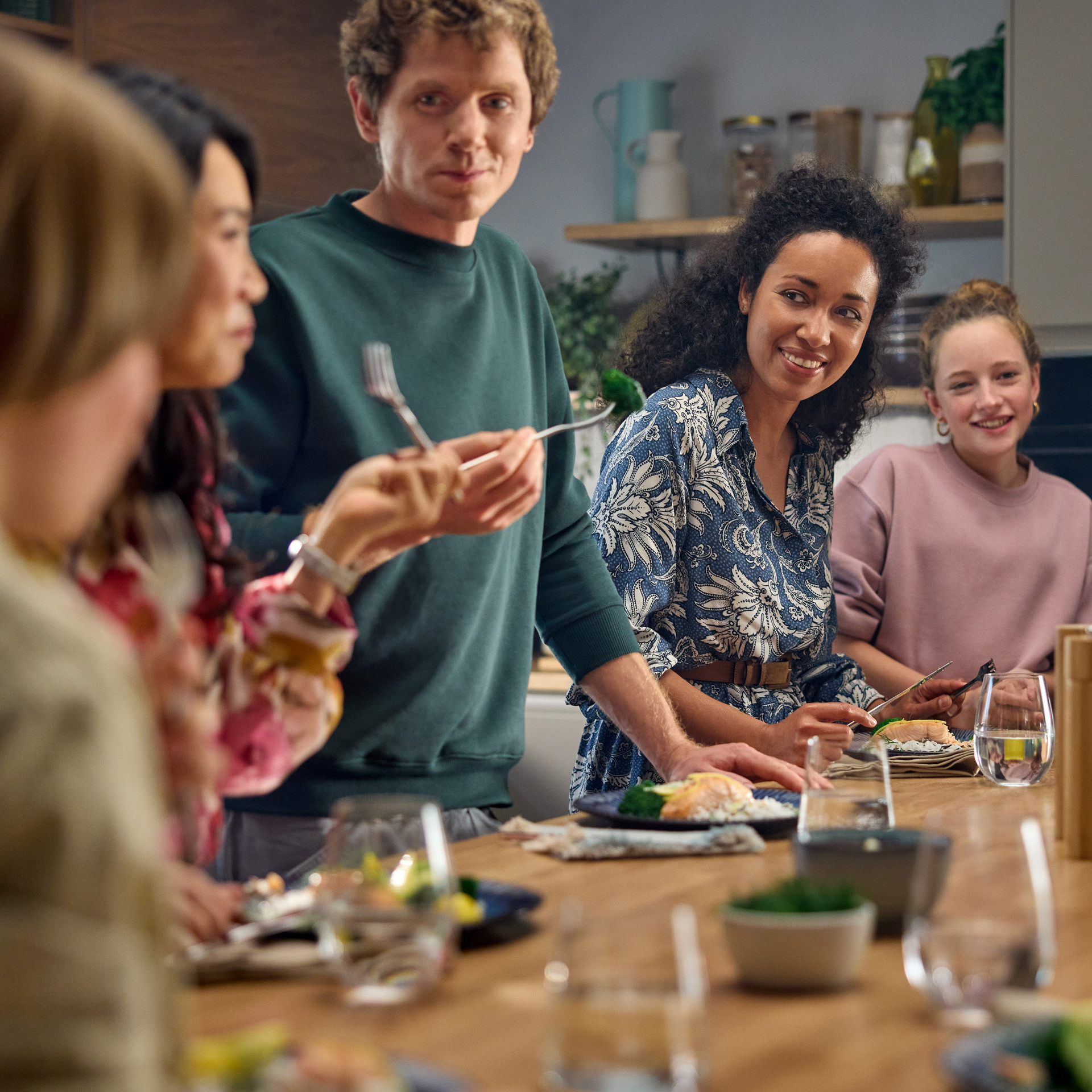 Gruppe von Personen teilt eine Mahlzeit an einem Küchentisch, Besteck und Gläser sichtbar.