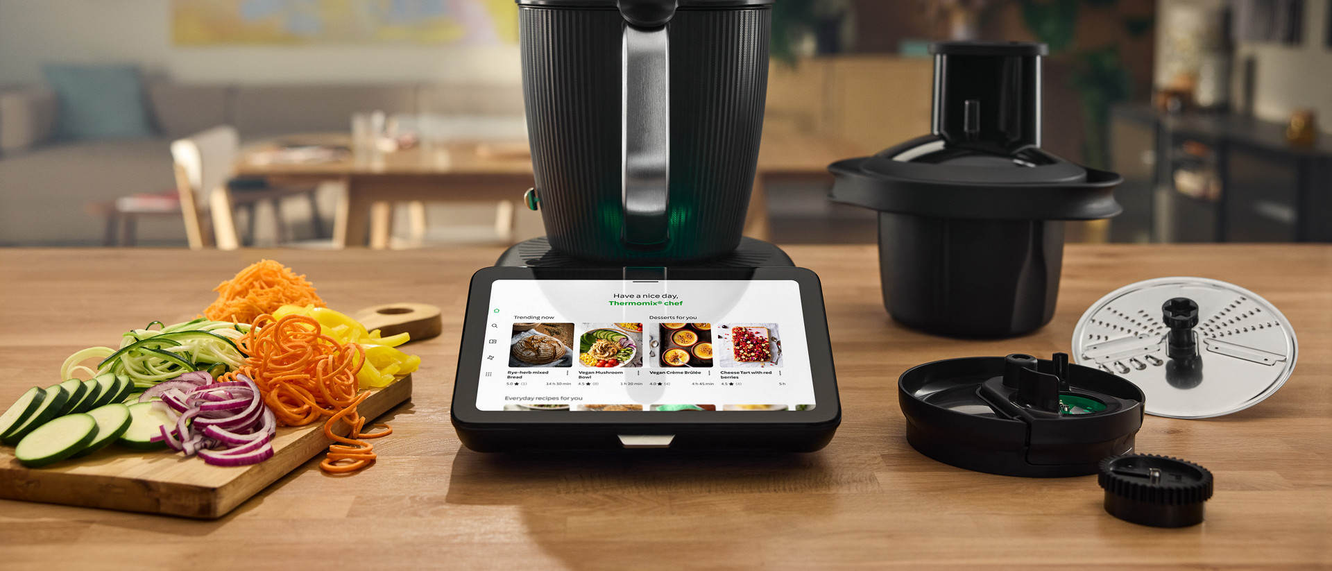 Thermomix® TM7 with Cutter Plus accessories on a kitchen table, surrounded by sliced vegetables and cooking tools.