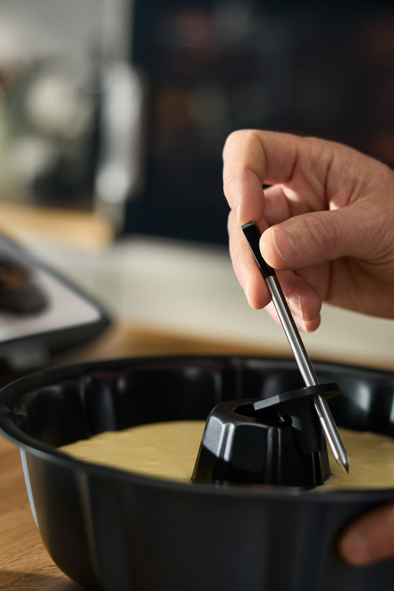 Person steckt einen Thermomix® Sensor in Kuchenteig in einer Backform, Anleitung auf einem Tablet sichtbar.