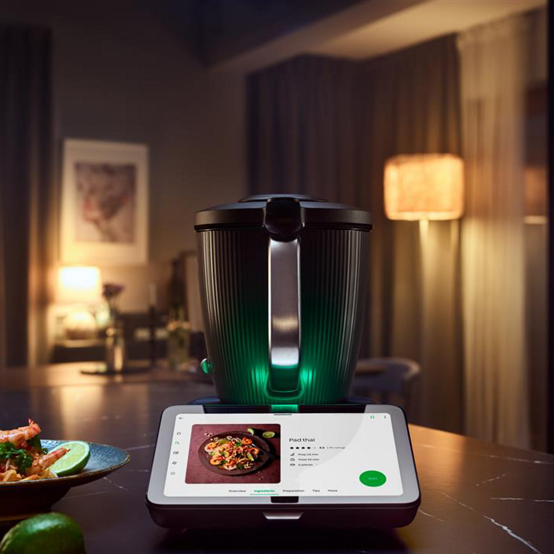 Thermomix® TM7 on a kitchen table, touch display showing a Pad Thai recipe, surrounded by food ingredients.