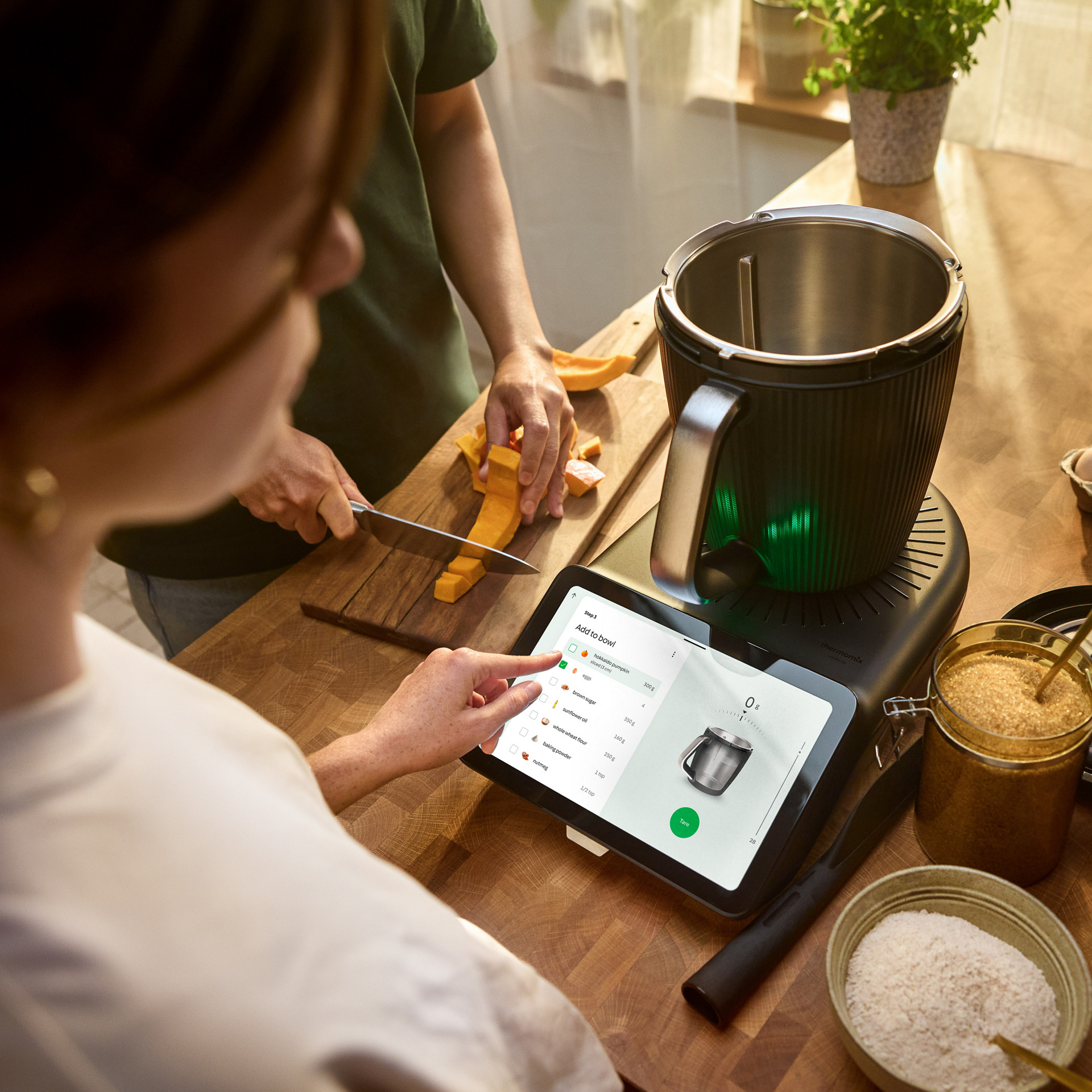 Two people cooking together using the Thermomix® TM7, following a recipe on the touchscreen, with ingredients on the table.