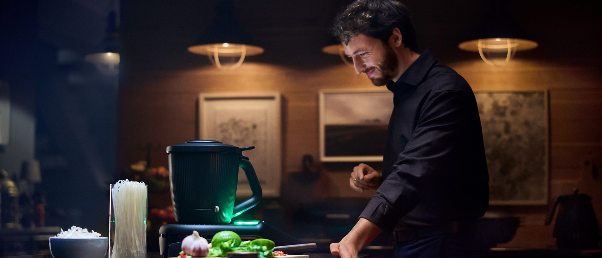 Person in einer Küche neben einem Thermomix® TM7 mit Kochzutaten auf der Arbeitsplatte bei warmem Licht.