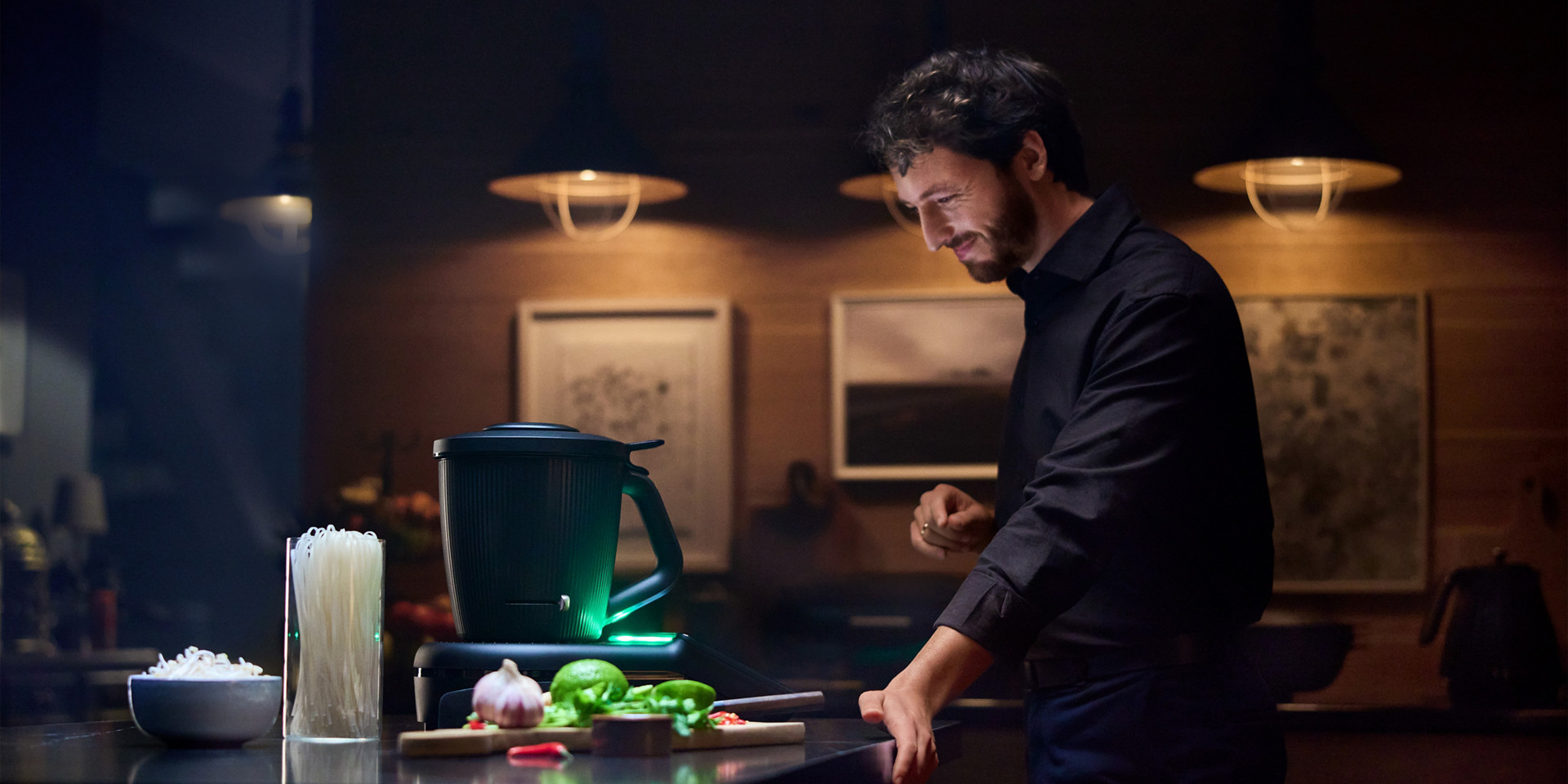 Persona en una cocina junto a un Thermomix® TM7 con ingredientes para cocinar en la encimera, bajo luz cálida.