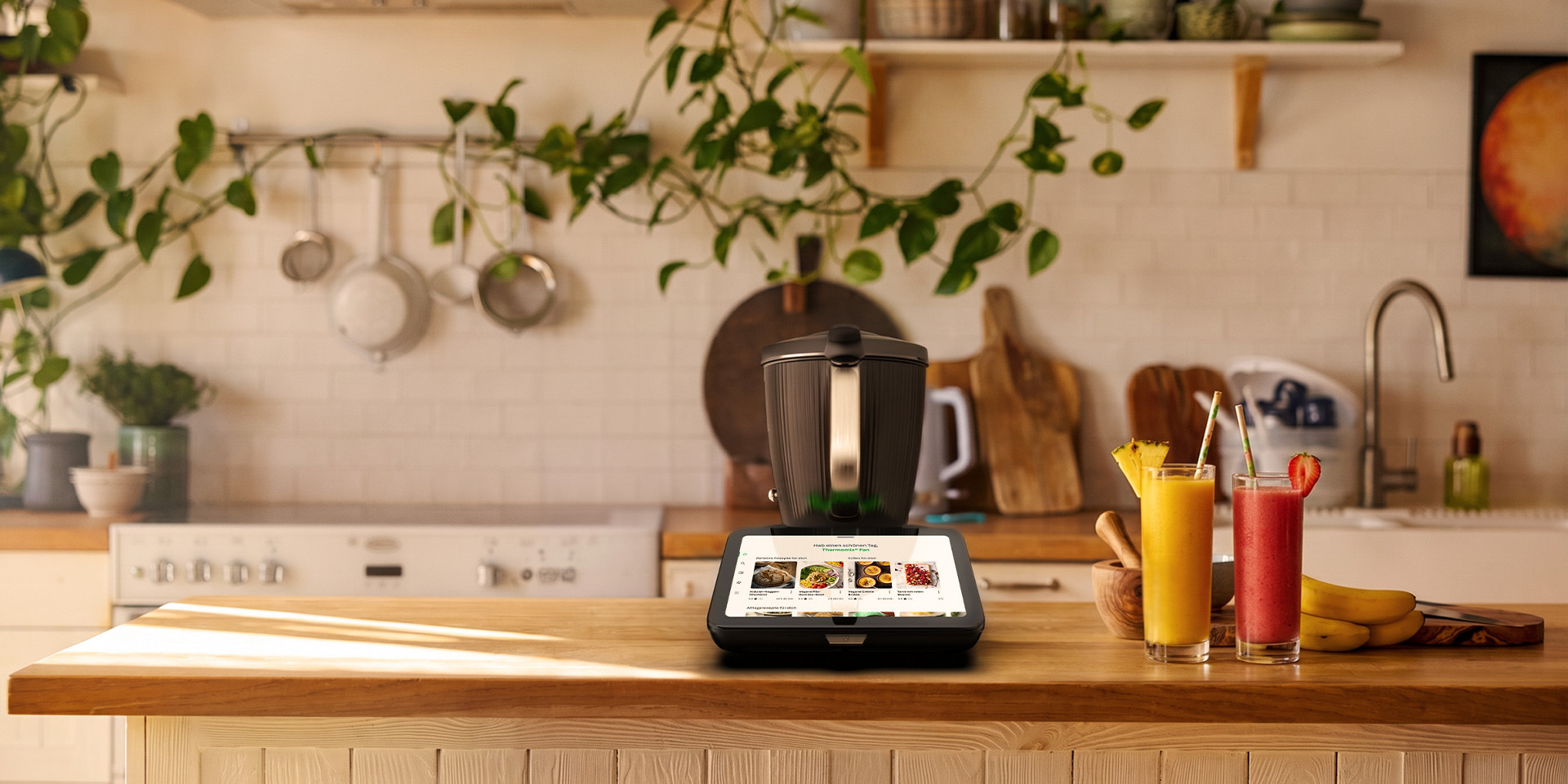 Thermomix® TM7 steht auf einer Küchenarbeitsfläche mit zwei Smoothies; Küchenutensilien und Pflanzen im Hintergrund.