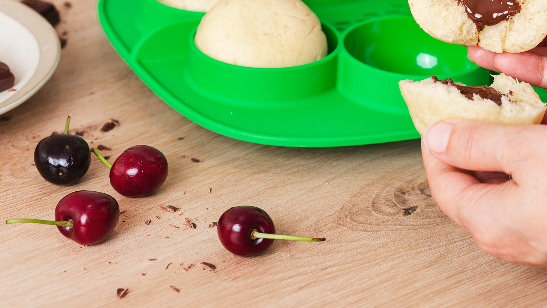 Osoba trzyma bułkę z czekoladą nad zieloną formą, w tle wiśnie, czekolada i Thermomix® TM7.