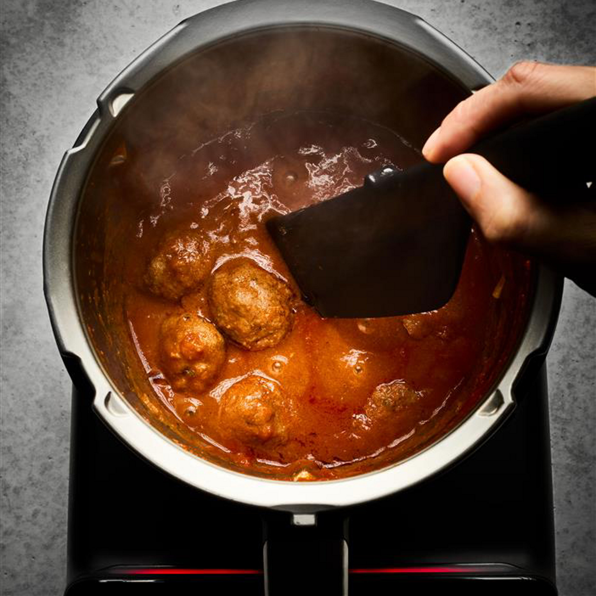 Hand stirring sauce inside an open Thermomix® TM7 mixing bowl, steam rising in a kitchen setting.