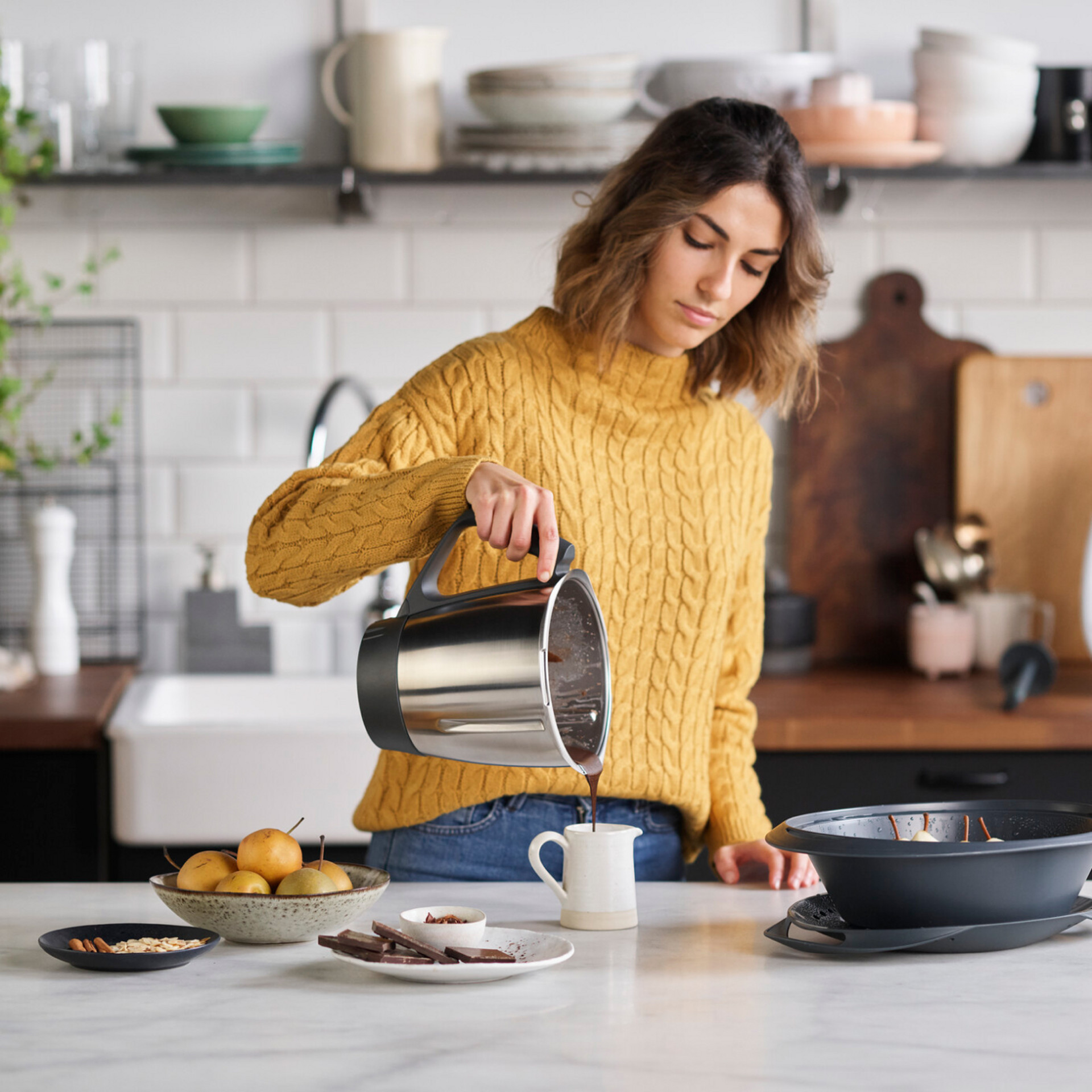 Mujer vierte chocolate del bol de Thermomix® TM6 en una taza en la cocina, rodeada de fruta y chocolate.