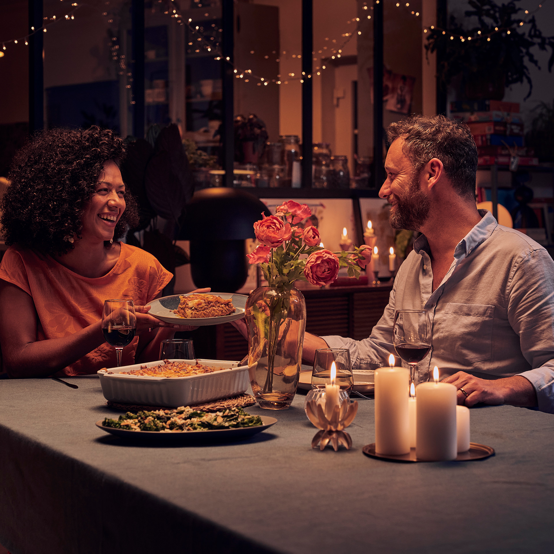 Two people share a meal at a candlelit dinner table with flowers and dishes in a cozy, warmly lit home setting.