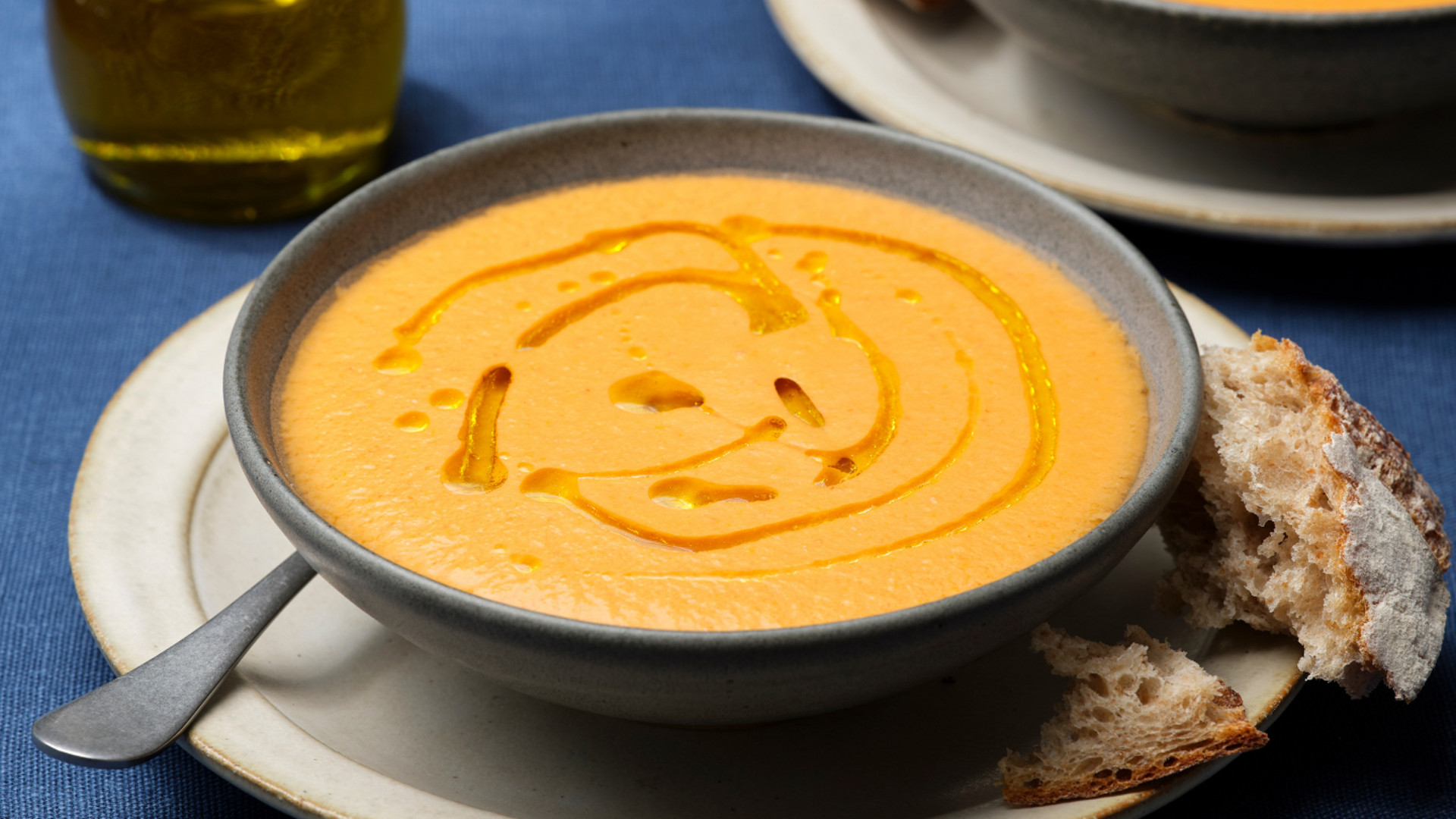Sopa cremosa alaranjada com fio de azeite em tigela cinza, servida com pão rústico num prato.