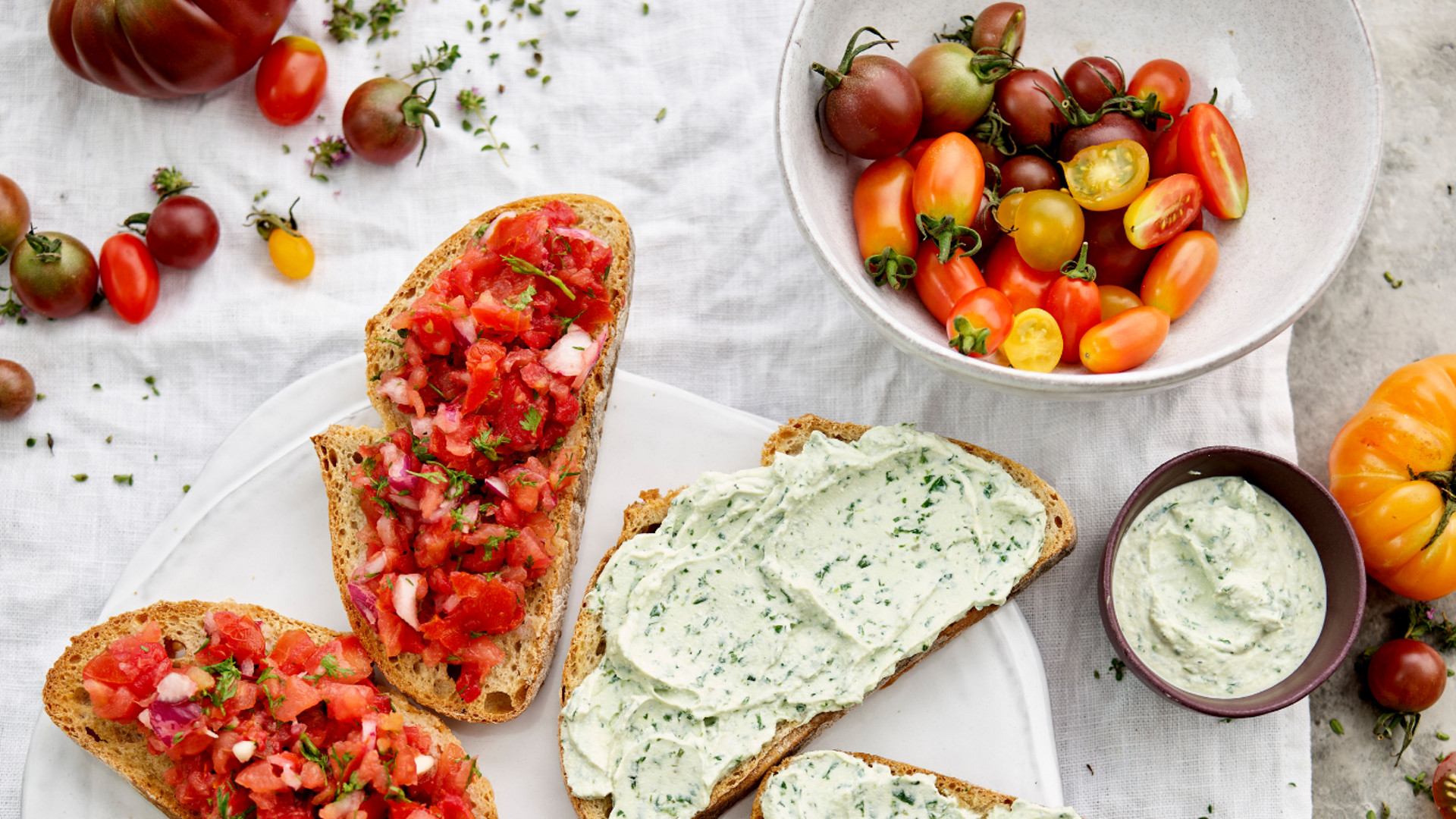 Fatias de pão com bruschetta de tomate e queijo-creme de ervas, taça de tomates coloridos e taça de creme de ervas sobre toalha branca.