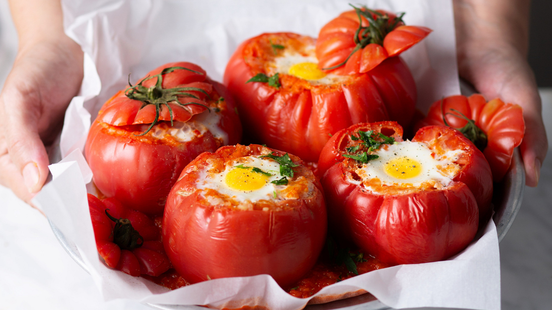 Mãos seguram uma travessa com tomates assados recheados com ovos e ervas sobre papel vegetal branco.