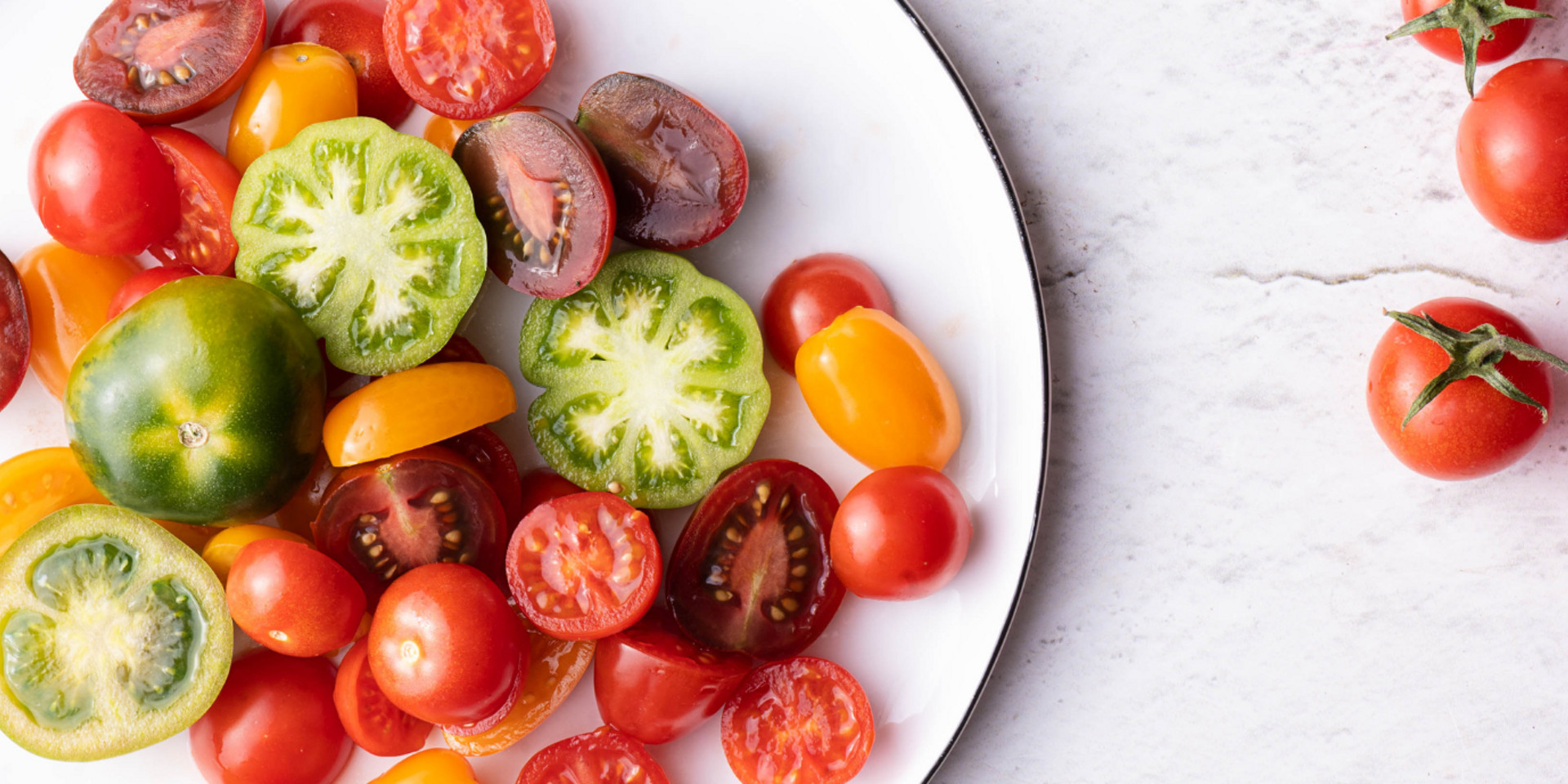 Tomates variados inteiros e fatiados de várias cores num prato branco, com mais tomates em mármore claro.