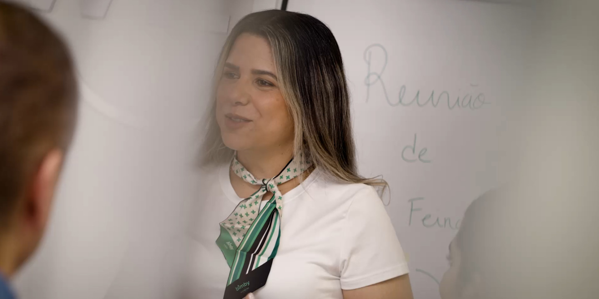 Mulher com lenço verde Bimby conduz reunião; “Reunião de Fena” escrito num quadro branco ao fundo.