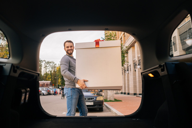 Delivery man holding large festive box with red bow near car outdoors city street and looking at camera. Courier male delivering gift to client. Shooting from trunk of car.