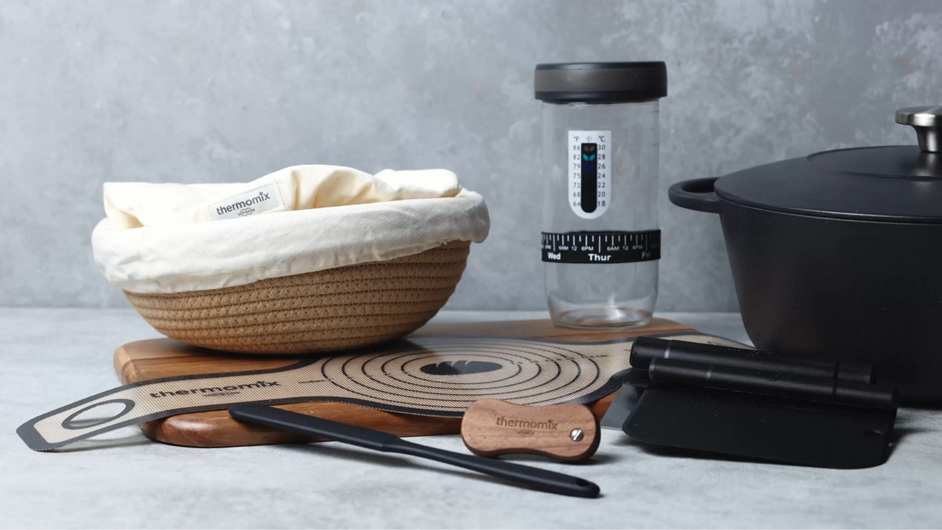 Thermomix® Backzubehör mit Gärkorb, Silikonmatte, Glas, Spatel und Teigwerkzeugen auf einer Arbeitsfläche.