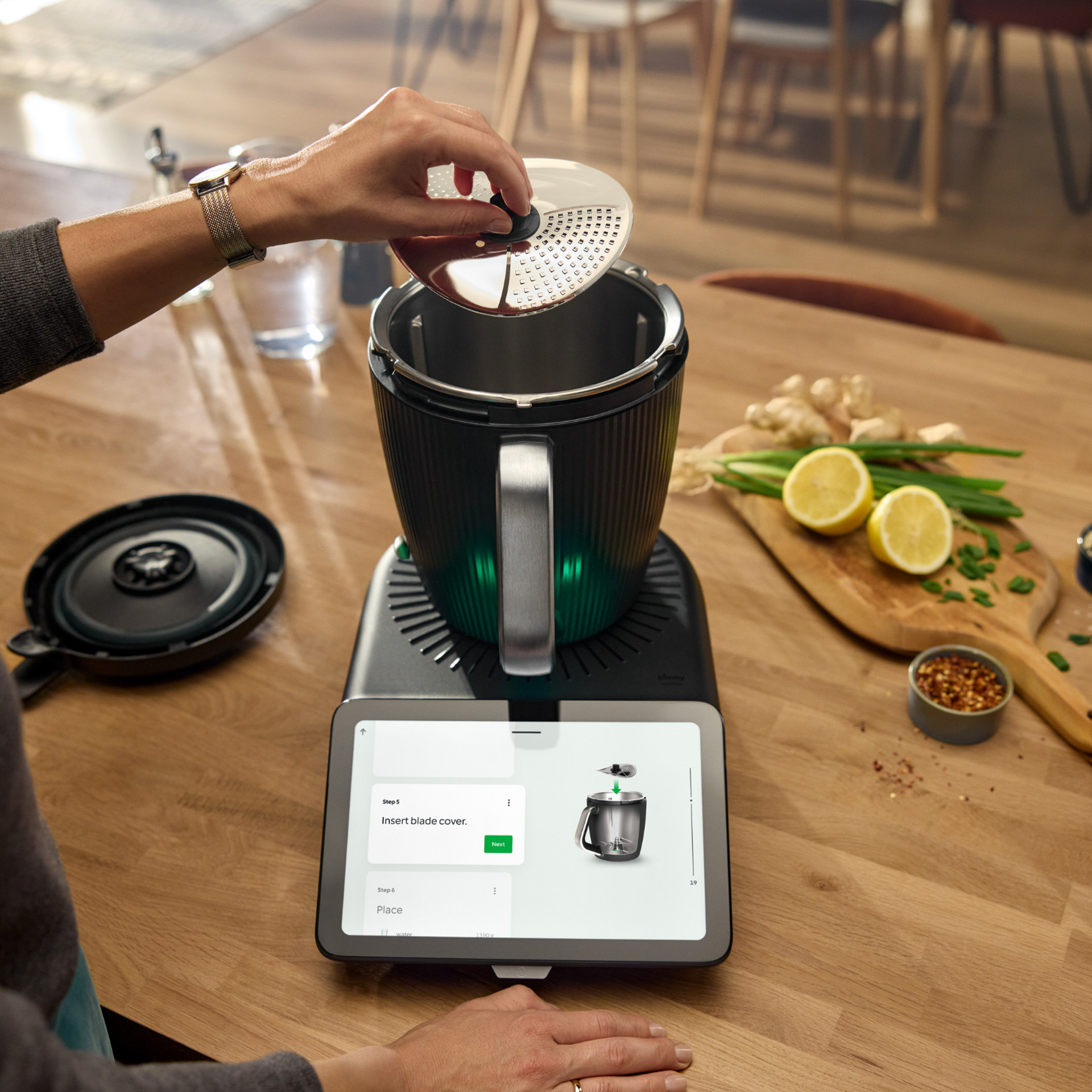 Pessoa insere cobertura de lâmina no Bimby® TM7, tablet com instruções, ingredientes sobre a mesa.
