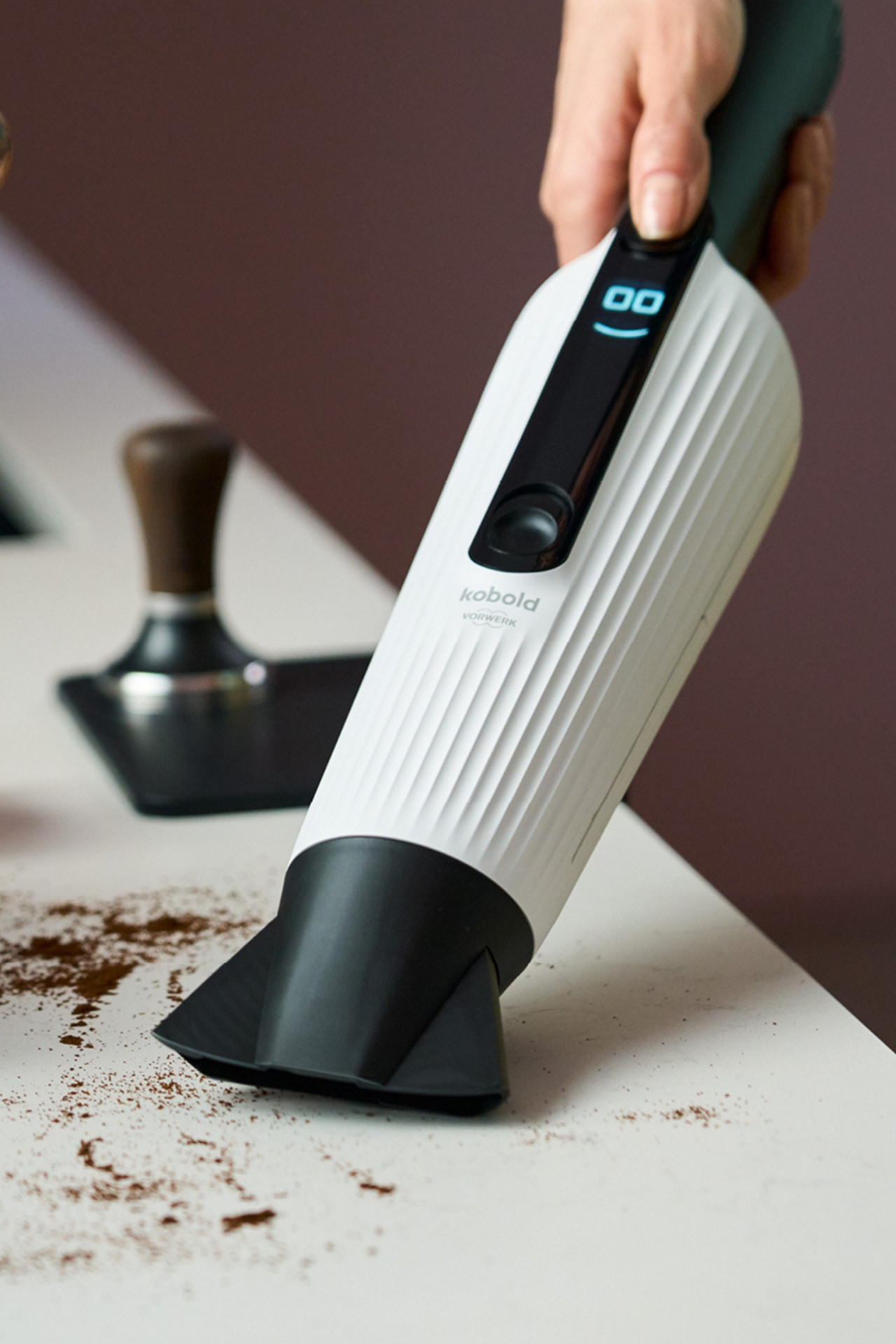 Mão utiliza Kobold VM7 Aspirador de Mão para limpar pó de café numa bancada, junto a um compactador de café.