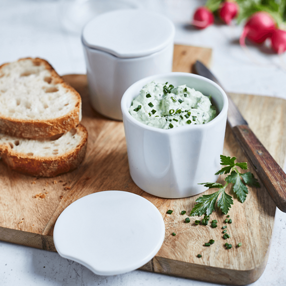 Fatias de pão e recipientes de cerâmica com queijo creme de ervas numa tábua de madeira decorado com salsa e cebolinho