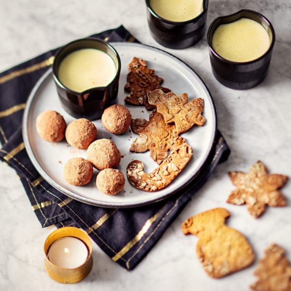 Prato com bolachas decoradas trufas de chocolate três chávenas de bebida cremosa vela e guardanapo sobre mesa de mármore