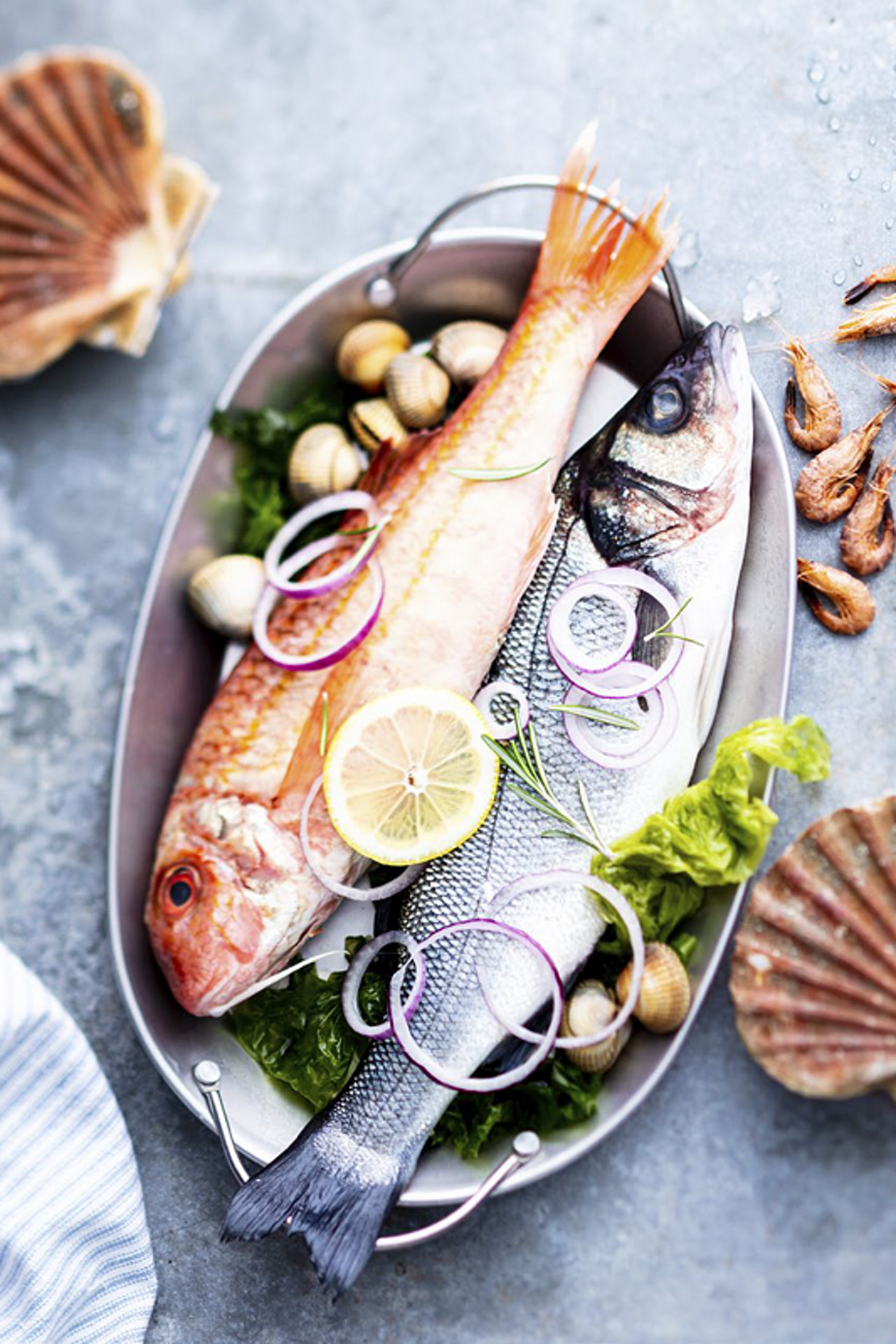 Two raw fish in a tray with lemon slices, onion rings, shellfish, lettuce, and shrimp on a stone surface.