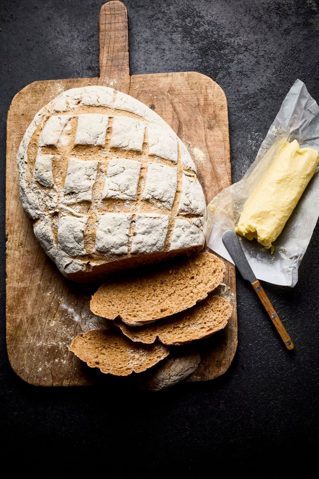 Rustic loaf of bread with three slices on a wooden board, next to a stick of butter and a butter knife.