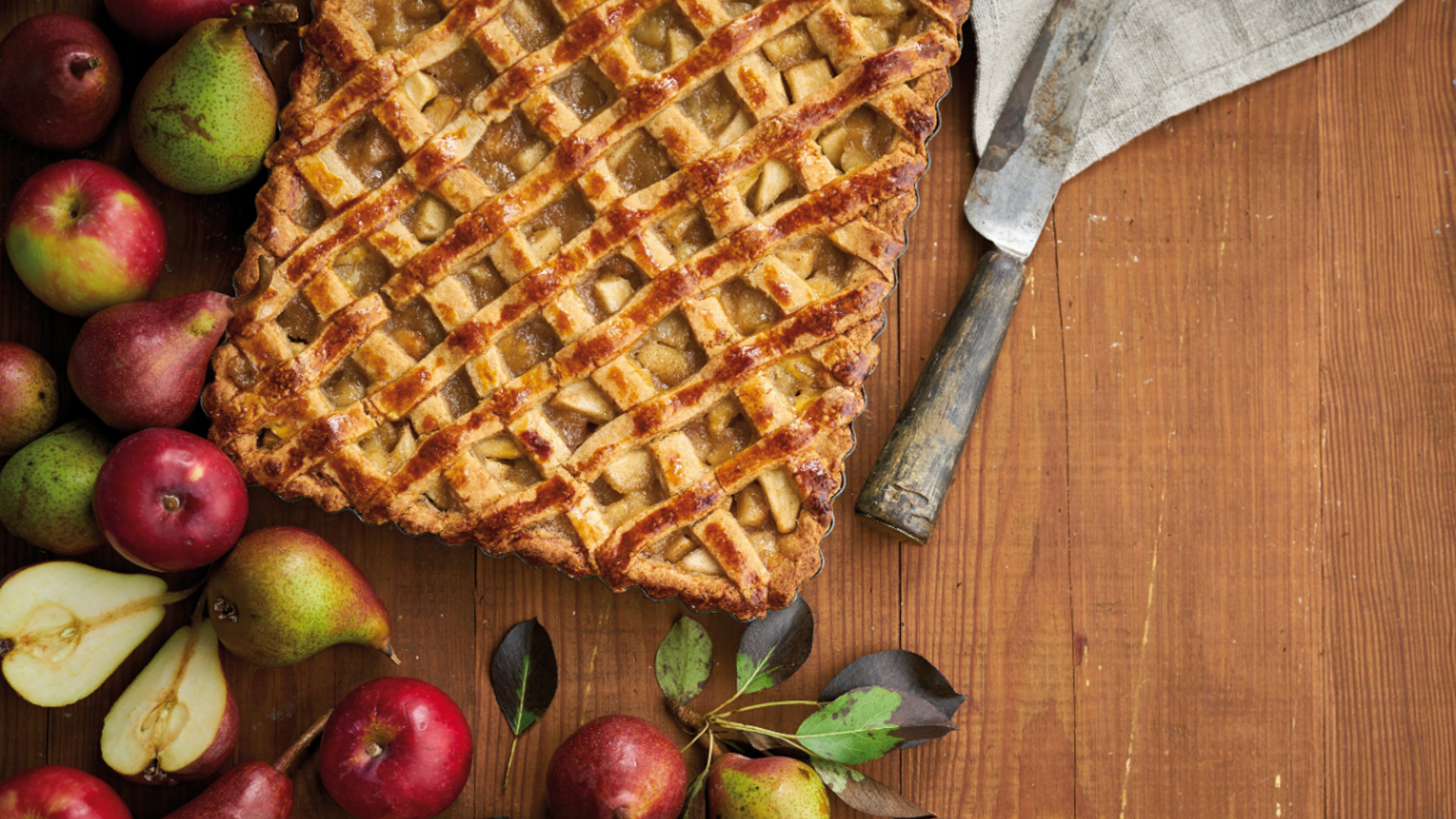 Tarte retangular de maçã e pêra com cobertura de rede, em mesa de madeira, rodeada de maçãs e peras, com espátula ao lado.