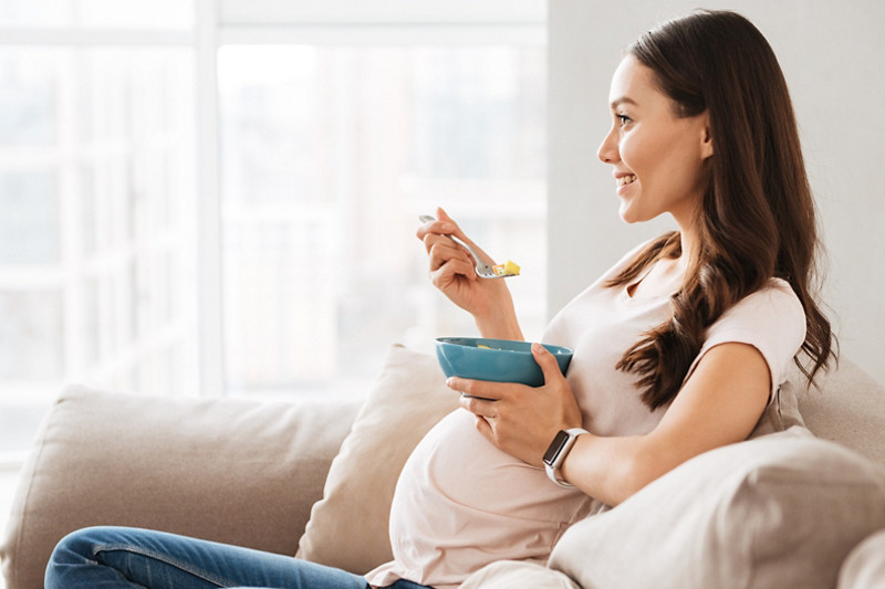 Smiling pregnant young woman having healthy breakfast while sitting on a couch