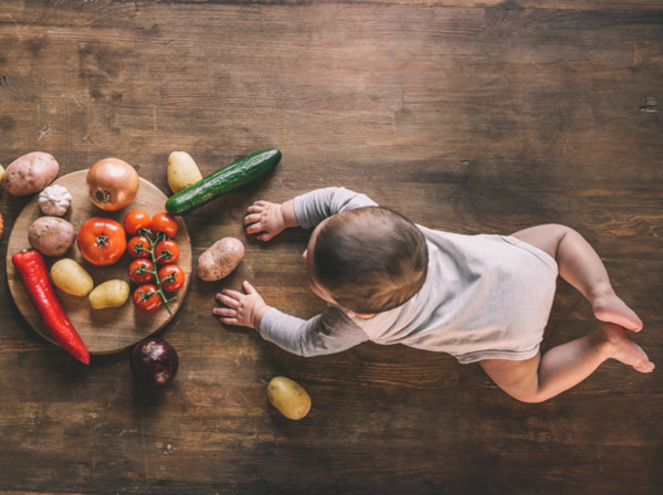 Overhead view of cute baby boy lying on kitchen table near group of vegetables on chopping board