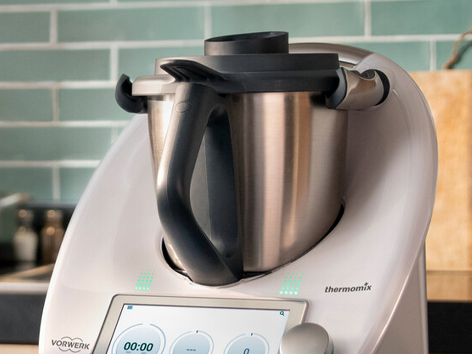 Thermomix® TM6 auf einer Küchenarbeitsplatte, mit gefliester Wand und einem Holzbrett im Hintergrund.