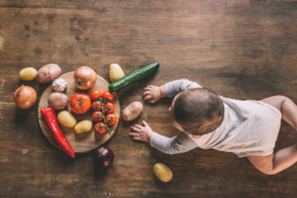 Overhead view of cute baby boy lying on kitchen table near group of vegetables on chopping board