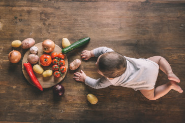 Overhead view of cute baby boy lying on kitchen table near group of vegetables on chopping board