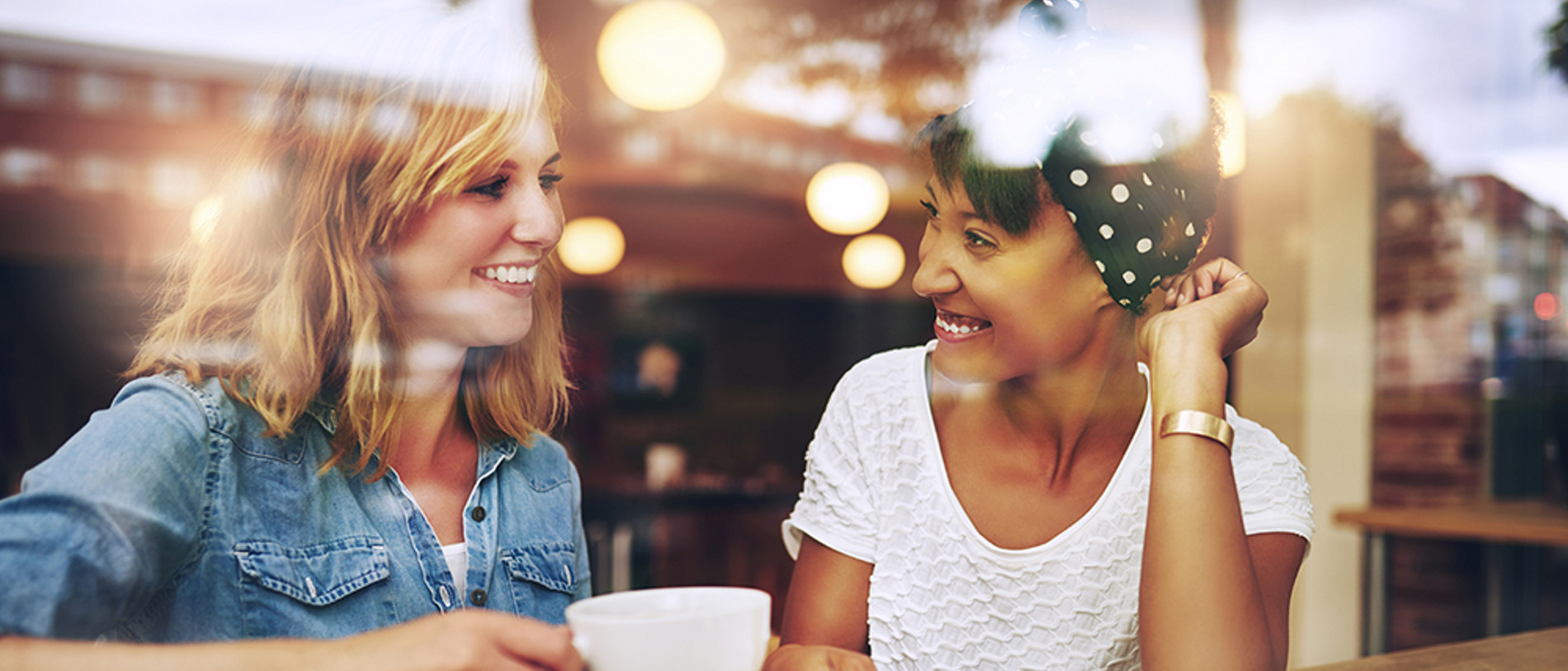 Two multi ethnic friends enjoying coffee together in a coffee shop viewed through glass with reflections as they sit at a table chatting and laughing