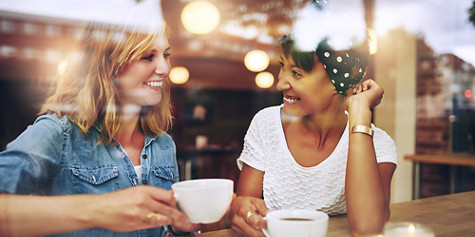 Two multi ethnic friends enjoying coffee together in a coffee shop viewed through glass with reflections as they sit at a table chatting and laughing